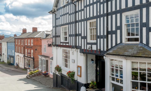 A view of the front of the Dragon Hotel with a row of colorful buildings down Broad Street, including a black and white timber-framed structure, under a partly cloudy sky.